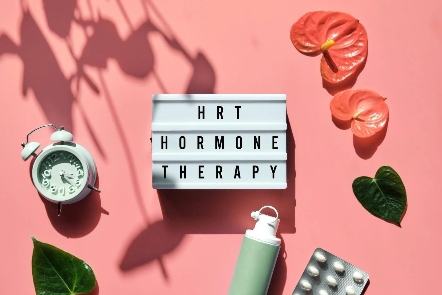 A flat lay of a lightbox displaying "HRT HORMONE THERAPY," with an alarm clock, plant leaves, and medication on a pink background.