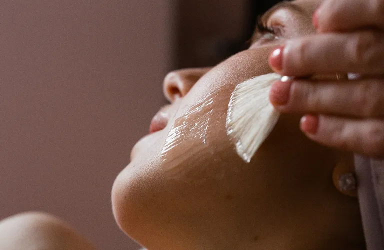 Close-up of a person receiving a facial treatment with a brush applying a clear gel on the cheek.