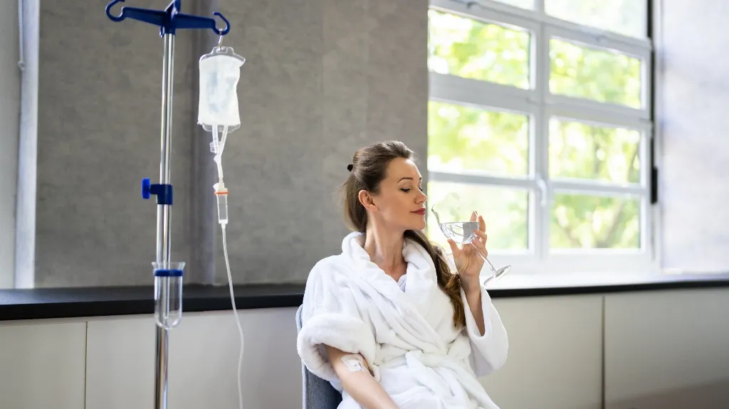 Patient in white robe sitting next to IV drip, holding a steaming glass in a bright room.