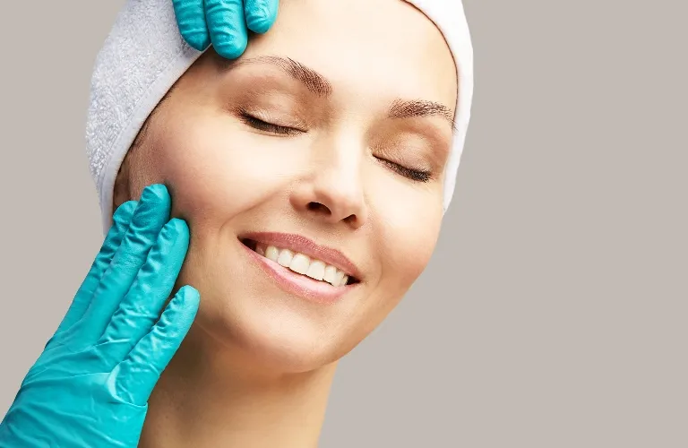 Close-up of a smiling woman with closed eyes receiving a facial treatment, wearing a white headband, with blue-gloved hands gently touching her face.