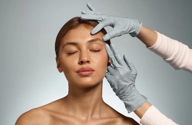 Close-up of a woman with closed eyes receiving a facial examination from a person wearing gray gloves.