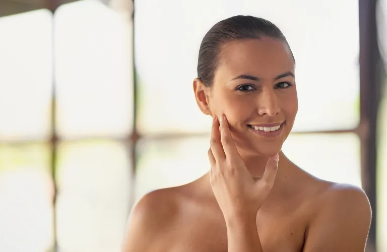 Portrait of a smiling woman with clear skin touching her face against a bright window background.
