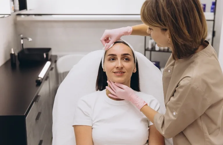 A skincare professional wearing pink gloves examines a seated woman's face in a clinical setting.