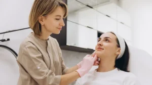 Medical professional in beige uniform administering facial injection to seated patient in clinic.