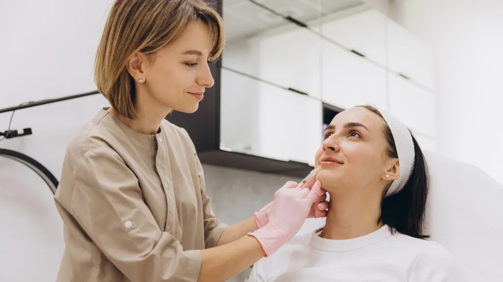 Medical professional in beige uniform administering facial injection to seated patient in clinic.