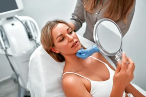 Woman in white tank top examining her face in a handheld mirror during skincare treatment.