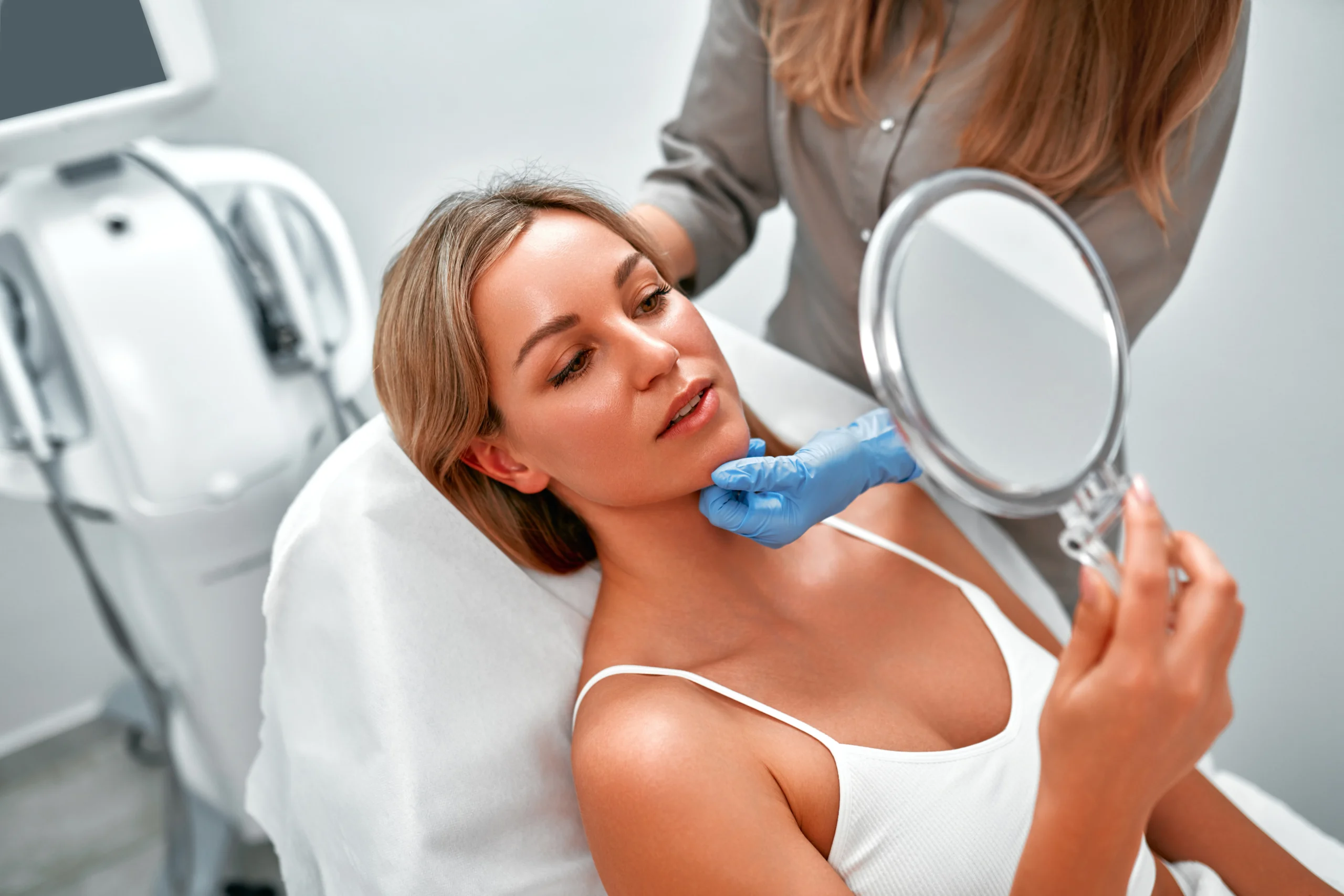 Woman in white tank top examining her face in a handheld mirror during skincare treatment.